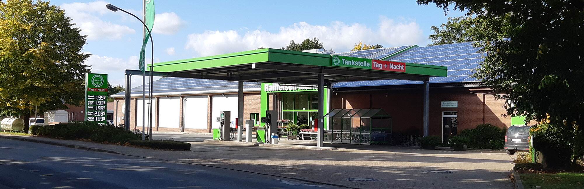 Du siehst eine RAISA Tankstelle mit grünem Vordach, Solaranlage und Preistafel an einem sonnigen Tag.